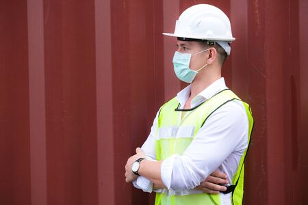 American engineers workers wear a safety vest and mask to prevent coronavirus. Standing arms crossed in front of the container at the container shipping area. Industrial and COVID protection concept.の写真素材