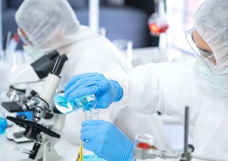 Young Asia Scientist in virus protection suit pouring blue solution from cylinder to the flask in the laboratory. There is a microscope and biohazard sign on the flask on the table. Research concept.の写真素材