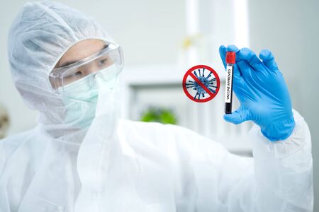 Scientists wear virus protection cloth and hold COVID 19 vaccine samples on his hand with coronavirus sign and blurred background. He looks at the test tube and success to win coronavirusの写真素材