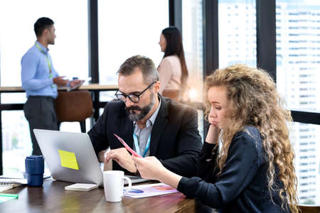 Two American people sitting at the table in front of the laptop and feeling boring at work. Unsuccessful of business. Women holding a file and feeling tired. Messy of table. Business concept.の写真素材