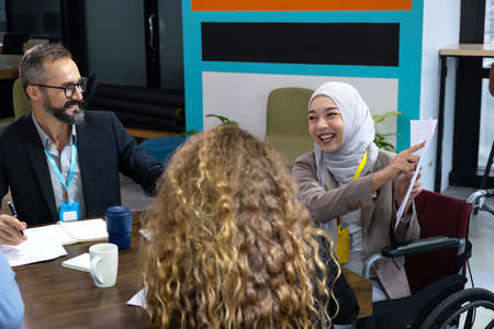 Muslim women sit in a wheelchair and present the trend of the market to multicultural working team. Feeling smile with her work. Team of business of different ethnicity, Caucasian, Asian.の写真素材