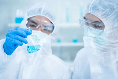 Two young Scientists in virus protection suit and wear hygiene mask seeing blue solution in the flask in the laboratory. Research on coronavirus protection. Research and education concept.の写真素材