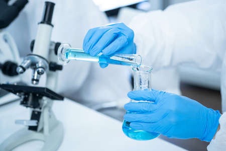 Young Scientists in virus protection suit pouring blue solution from cylinder to the flask in the...