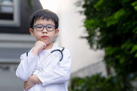 Asia boy wearing a medical suit and place a doctor stethoscope on his neck standing in the garden under sunlight. A boy crossed arm and touch his chin. Feeling happy and confident.の写真素材