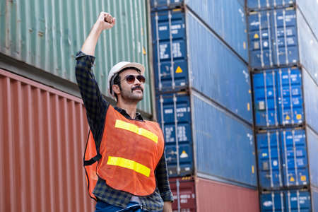 A confident engineer or technician wears a white helmet. Standing at the container shipping area. Raises his hand and proud to succeed in work. Feeling happy and fun. Industrial and shipping concept.の写真素材