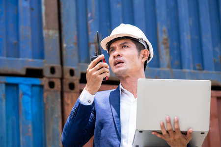 Businessman wearing safety helmet and holding a laptop, using radio communication in the container yard shipping area. Talking to the worker. Feeling stress. Industrial and shipping business conceptの写真素材