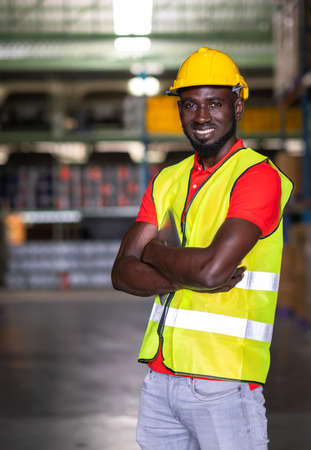 The engineer worker holds tablet crossed arm and standing in the warehouse distribution center. Man wears a safety helmet and vest. In background shelves with goods. Feeling smile and happy workの写真素材