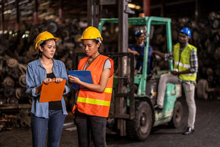 Engineer woman wearing helmet standing at the large warehouse where keep machine parts and holding...