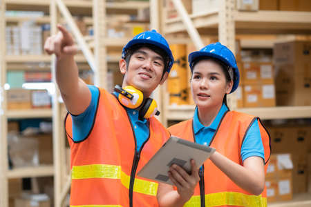 Man worker holds a tablet and points at shelves and lets women looking at the parcel in the large warehouse store.  Engineers wear a helmet standing in the factory. In background shelves with goodsの写真素材