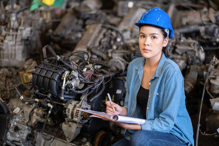 Confident Asia engineer woman worker wears a safety helmet takes note on the clipboard. look at the camera. Squat in the automotive spare parts warehouse. stock management concept. Many old enginesの写真素材