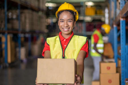 Smile woman workers carrying the box and reaching forward in the automotive parts warehouse distribution area. Female engineers people wear a safety helmet and vest. In background shelves with goodsの写真素材