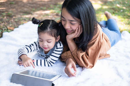 Closeup daughter with her mom using tablet and lying on the ground in the public garden. Girl playing game from the tablet. A good relationship at the weekend. Education, online and family conceptの写真素材