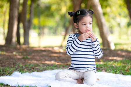Asia cute happy little toddle girl eating a soft bun (cake) and sitting in the public garden under sunlight with copy space. Outdoor activity. Food and nutrition for kid. Family and holiday conceptの写真素材