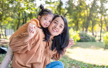 Asia daughter riding her mother back in the park with copy space. Family outdoor activity. Mom and kid smile and looking at the camera. Enjoy, happiness, and relationship concept.の写真素材
