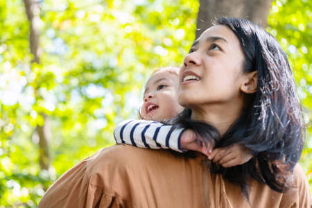 Asia daughter riding mother back and standing in the public garden or park under sunlight with copy...