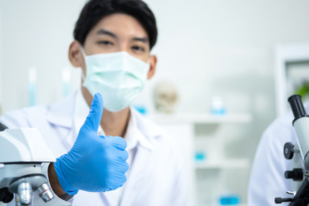 Scientists wearing hygiene masks, gloves, and lab coats sitting and thumb up in the laboratory. Research on the coronavirus vaccine to prevent COVID-19. Medical and health concept.の写真素材