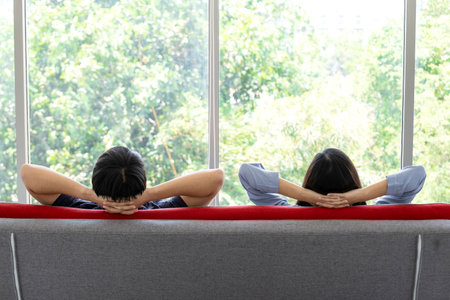 A man and woman sitting on the sofa in the living room and looking outside to see green background. Feeling relax and peaceful. Work from home. Quarantine concept.の写真素材