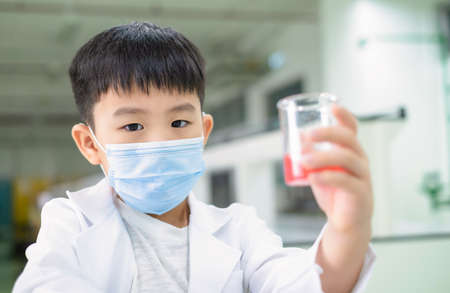 Portrait of a boy wearing hygiene mask and lab coat holding beaker which have pink solution in the laboratory. Kid looking forward and fun on experiment. Education, knowledge and learning conceptの写真素材
