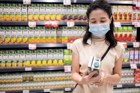 Asia woman wearing medical face mask and pick up orange juice from shelf. Reading nutritional value from the bottle. Shopping at supermarket in new normal style during coronavirus. Healthcare conceptの写真素材
