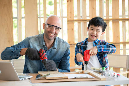 A senior carpenter and boy wearing goggle in the modern wood workplace and holding screwdriver. A...