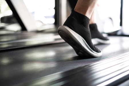 Close up of foot sneakers in the sport fitness gym. Man running on the track treadmill. Workout in the morning time. Routine exercise for health. Walking on belt with technology machine.の写真素材