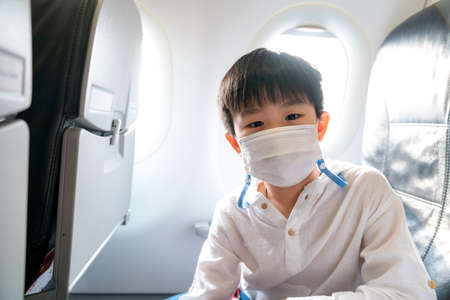 Asia boy wear face mask on the airplane and sit beside windows. Coronavirus disease pandemic outbreak impact to business airline. Travel during COVID 19 pandemic. New normal lifestyleの写真素材