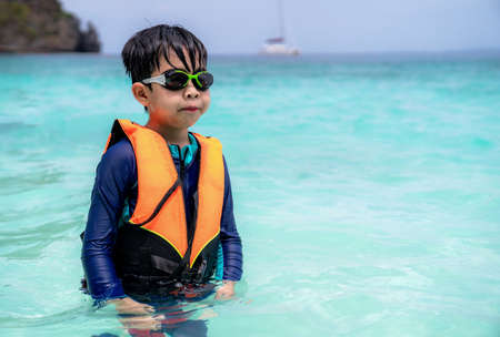 Asia child boy wearing goggles, swimming suit and life jacket standing in the sea at beach in Thailand with copy space. Summer vacation and holiday travel. Relaxation and happy in the waterの写真素材
