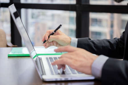 Businessman wearing suit holding pen and reading contract in the laptop before sign in the modern high office building. Success on the new project. Business concept and lifestyle in the cityの写真素材
