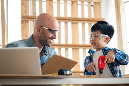 Professional carpenter wearing google and teach a boy use screwdriver in the modern wood workplace. Kid following teaching and point on the workpiece. Many tools and laptop on the table.の写真素材