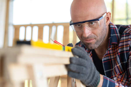 Senior carpenter wearing safety goggle and glove checking quality of wood and use spirit level for calculate in the wood workshop. High skill of workman. Building a furniture. Handcraft DIY concept.の写真素材