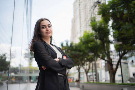 Confident professional business woman wearing suit and standing in front of office. Crossed arms and smile looking camera with blurred building background. Business, lifestyle and city life concept.の写真素材