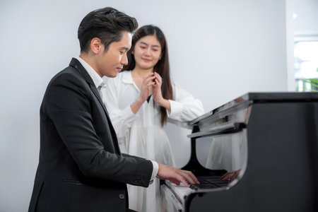 Pianist in black suit playing classic grand piano in the room. Professional musician young man happy and feeling relax. Pretty woman standing beside piano and clap. Practice before concertの写真素材