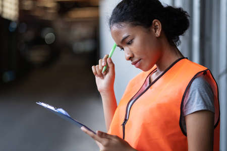 Africa American engineer woman wearing vest standing  at the automotive engine part in the factory. Worker holding clipboard and checking list in garage. Looking at note. Industrial and stock conceptの写真素材