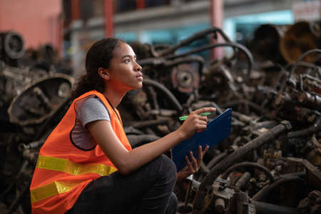 Africa American worker woman standing in the automotive engine part at the factory with copy space.  Holding clipboard and checking inventory of old engine in garage. Industrial and stock concept.の写真素材