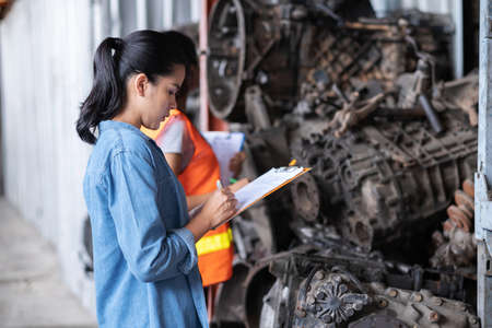 Asia engineer woman standing with Africa woman at the automotive engine part in the factory. Worker holding clipboard and checking list inventory of old engine in garage. Industrial and stock conceptの写真素材