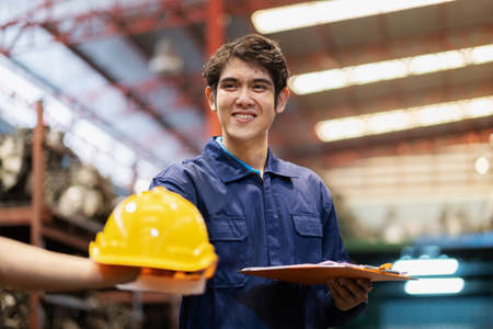Asia young engineer standing in the automotive part warehouse at factory. Received safety helmet and smile. Worker holding clipboard and check inventory of goods. Safety and stock conceptの写真素材