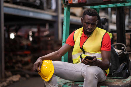 Africa American holding safety helmet and sitting at forklift in the automotive part warehouse. Worker look at smartphone and get the bad news. Feeling stress from fired from work. Disappointの写真素材