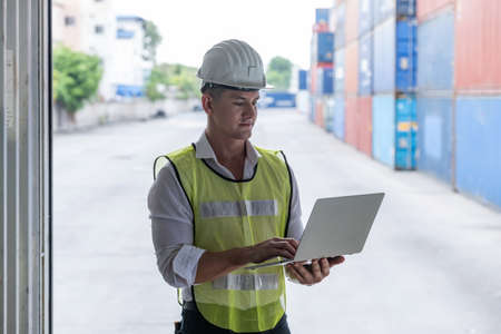 Confident of American engineer worker wearing helmet and vest standing in the container at shipping container terminal. Checking stock from laptop. Logistic, import or export concept with copy space.の写真素材