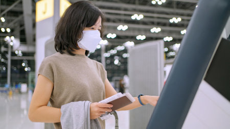 Woman wear mask using self service check-in kiosk at the international airport terminal. Touchscreen and see information on screen. Scan passport to record personal data. New normal travelの写真素材
