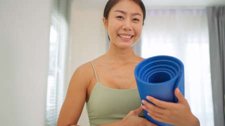 Close up of happy pretty woman wearing sportswoman bra holding yoga mat standing in the living room and smile. Healthy and active lifestyle. Refreshing after yoga practice. Exercise conceptの写真素材