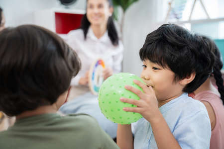 Handsome boy sitting in the kindergarten school classroom and holding green plastic ball and show to...