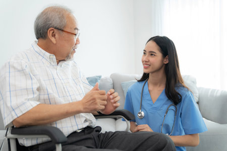Attractive caregiver nurse sitting beside elderly man who sitting on the wheelchair and smile at home. Doctor take care and encourage  patient. Rehabilitation and healthcare concept.の写真素材