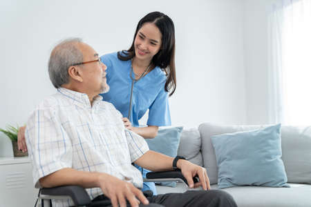 Attractive caregiver nurse stand behind elderly man who sitting on the wheelchair and smile at home. Doctor take care and encourage  patient. Rehabilitation and healthcare conceptの写真素材