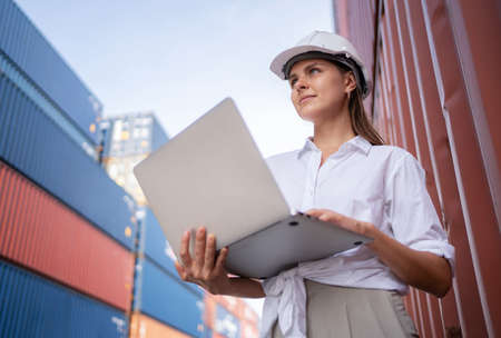 Dock manager or engineer worker in causual suit standing in shipping container yard holding laptop with smile. Import and export product. Manufacturing transportation and global business concept.の写真素材