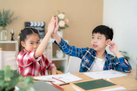Cute girl and boy sitting in the classroom and doing homework together. Doing hi five and looking at camera. Portrait of school kid. Learning, education and kindergarten concept. Back to schoolの写真素材