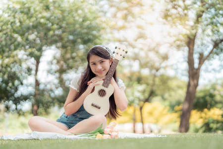 Pretty attractive woman sitting in the garden with blur background. Girl holding Ukulele Guitar on sunny day. Happiness during vacation. Relaxation, Aromatherapy freedom feeling lifestyleの写真素材