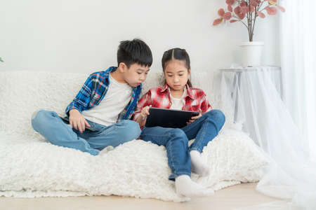 Portrait boy and girl sitting on the soda in the living room isolate on white background. Girl playing game in tablet and boy looking at tablet.  Feeling happy. Technology and learning conceptの写真素材