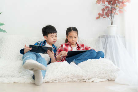Portrait boy and girl sitting on the soda in the living room isolate on white background. Girl playing game in tablet and boy looking at tablet.  Feeling happy. Technology and learning conceptの写真素材