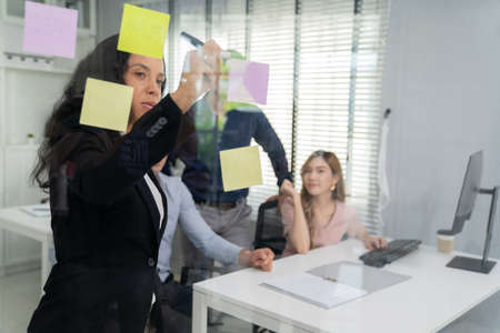 Executive businesswoman manager write some idea on sticky notes and put it on the glass board meeting with team. Managing professional organization business plan project presentation in officeの写真素材
