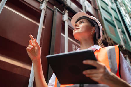 Dock manager or engineer worker in casual with vest standing in shipping container yard holding...